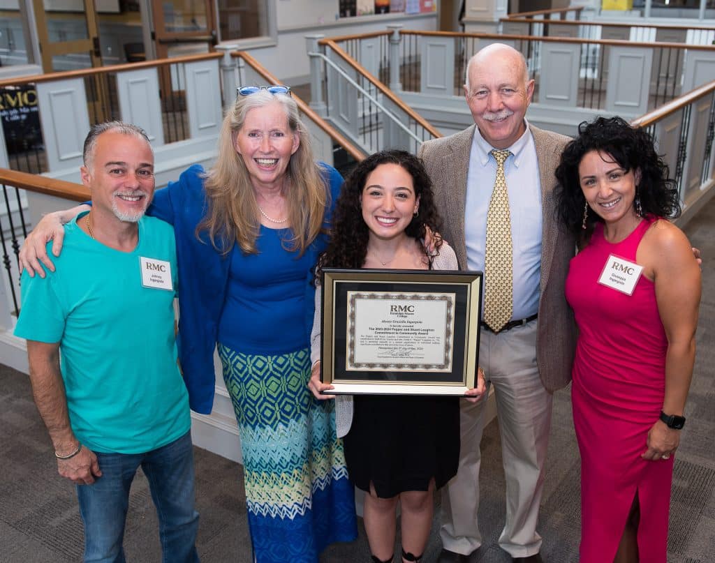 Five adults smiling in a lobby, a young woman in the center holding an award plaque, surrounded by four older adults, two women and two men, in casual and business attire.