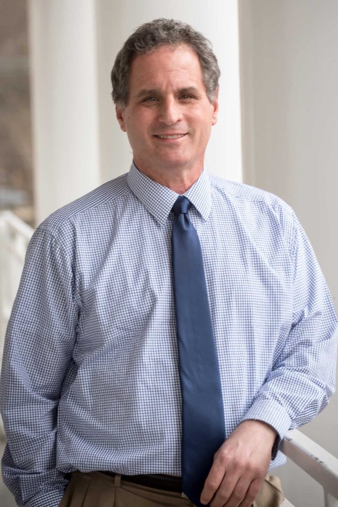 A man in a checkered dress shirt and navy tie stands, leaning slightly on a railing, with a neutral expression and a background of white columns.