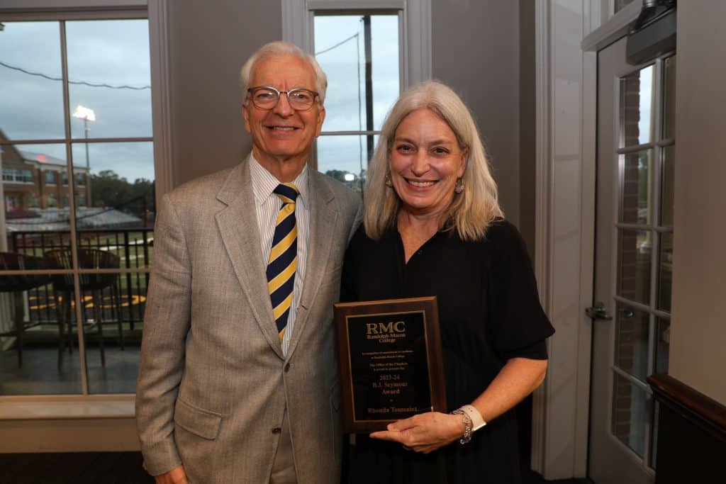 An older man in a suit and an older woman in a black dress smiling, holding a commemorative plaque at an indoor event.