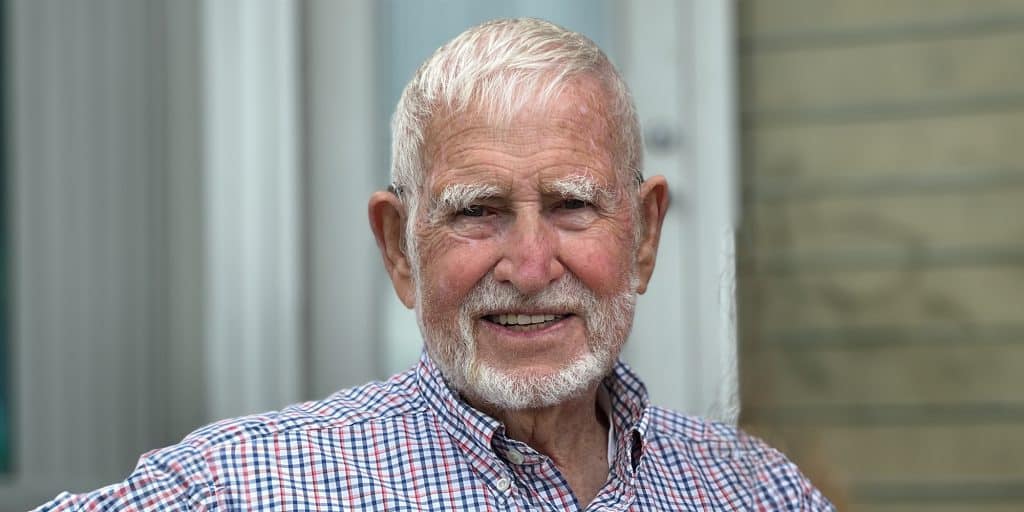 Elderly man with white hair and beard, wearing a checkered shirt, smiling in front of a blurred background.