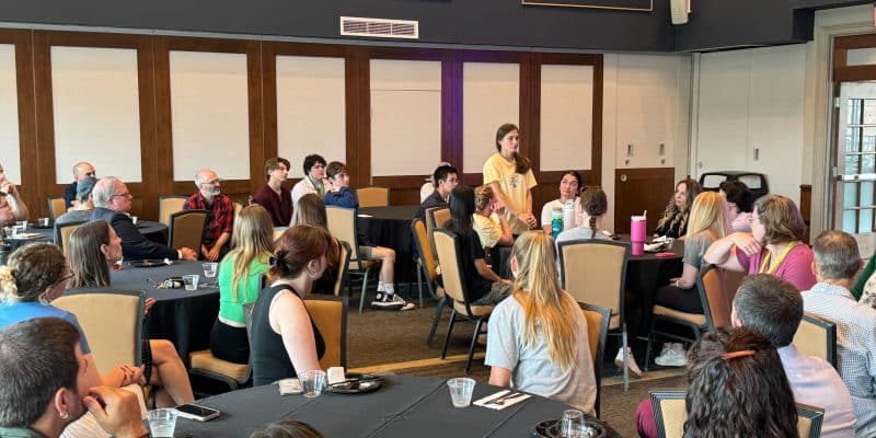 A group of people sitting at round tables in a conference room, listening to a person standing and speaking near the front.
