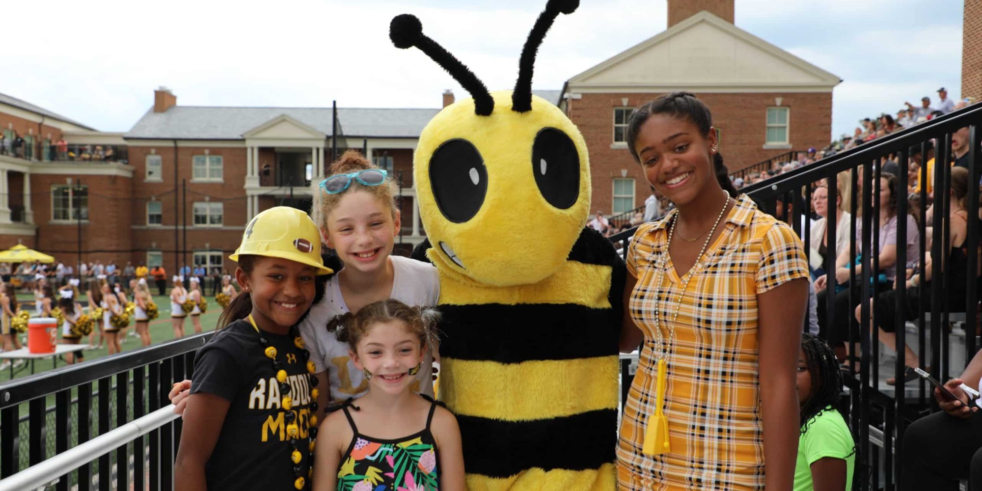 Four children smile while posing with a person in a bee mascot costume at an outdoor event. Dubbed the Buzz Buddies, this group of friends enjoys the lively atmosphere, with a marching band and spectators visible in the background.
