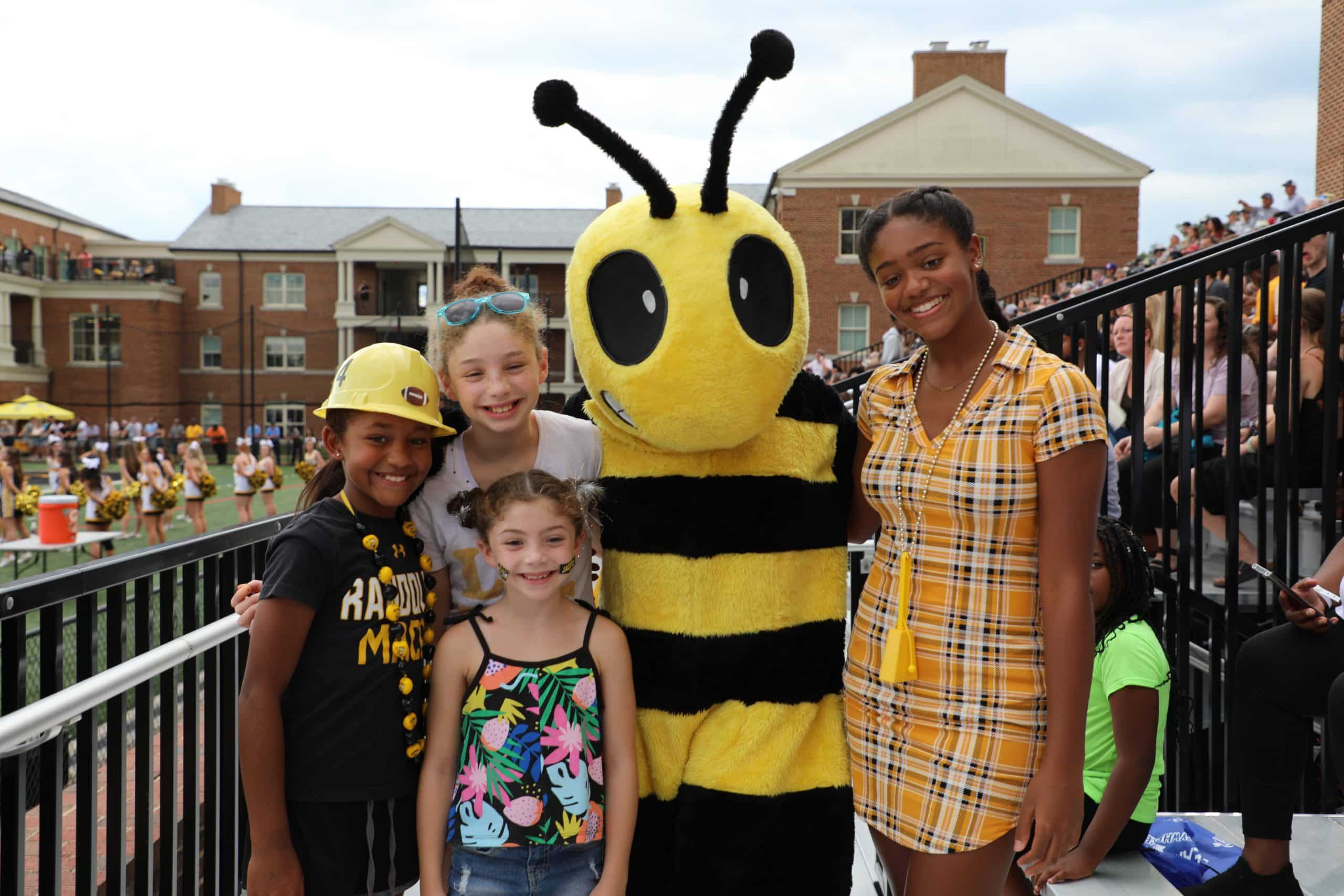 Four children smile while posing with a person in a bee mascot costume at an outdoor event. Dubbed the Buzz Buddies, this group of friends enjoys the lively atmosphere, with a marching band and spectators visible in the background.