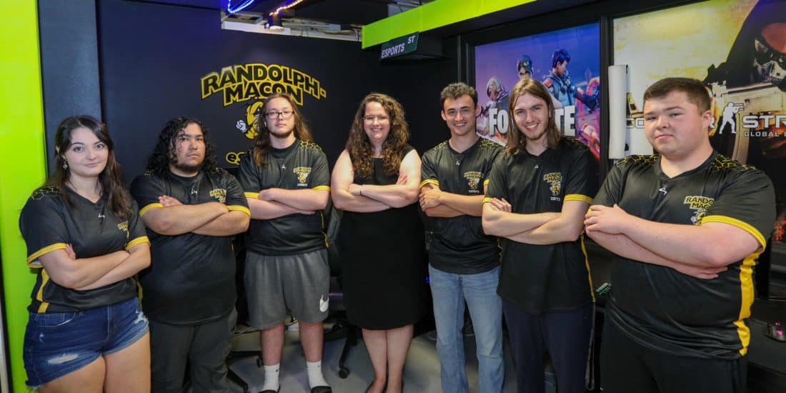 A group of people in black uniforms with yellow accents stand with arms crossed, posing in front of a "Randolph-Macon" sign and several posters in a room with neon lighting.