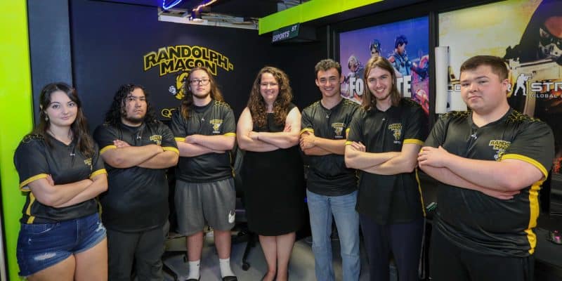 A group of people in black uniforms with yellow accents stand with arms crossed, posing in front of a "Randolph-Macon" sign and several posters in a room with neon lighting.