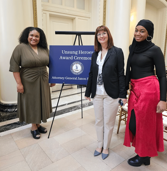 Three women standing in front of a sign that reads "Unsung Heroes Award Ceremony." at an event organized by the Attorney General.