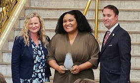 Three people are standing on stairs. The person in the center is holding an award. The person on the left and right are smiling. A gold railing is visible in the background.
