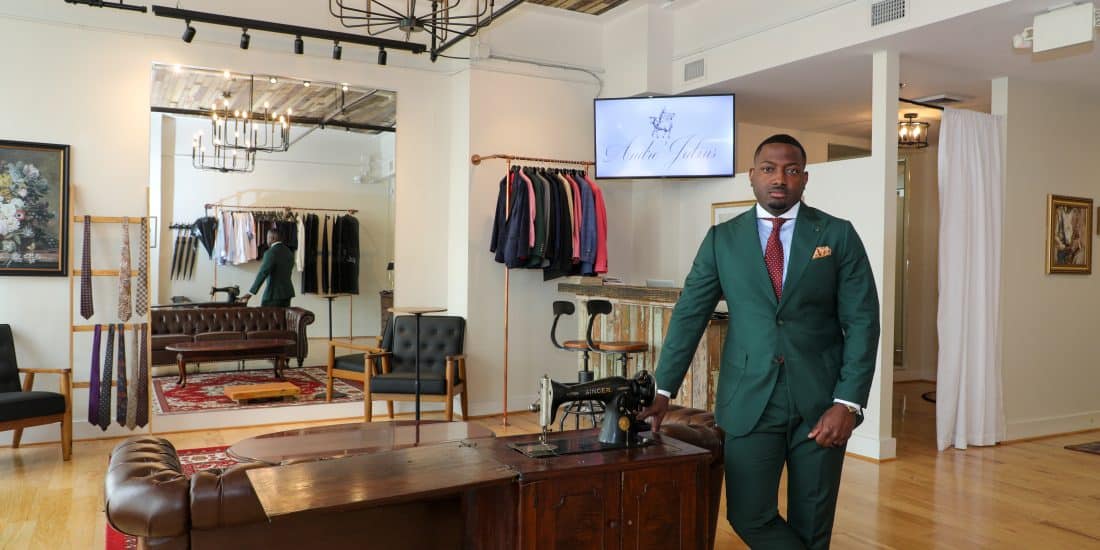 A man in a green suit stands beside a wooden sewing machine table in a tailor shop with suits and accessories displayed in the background.