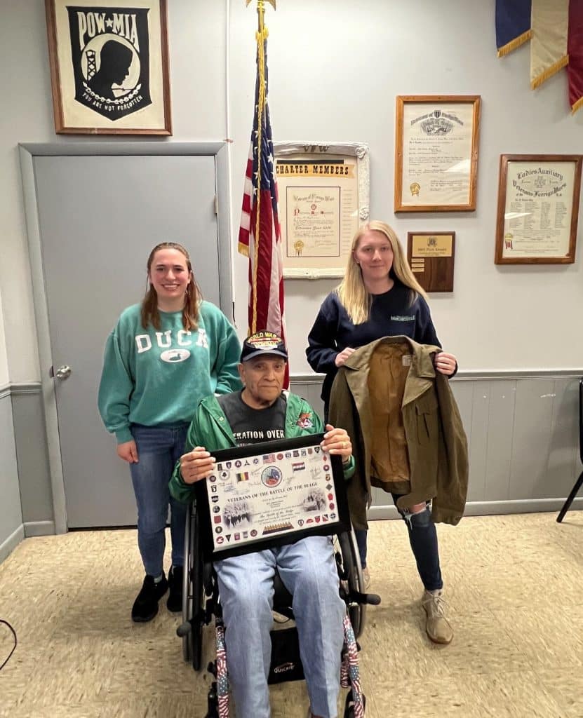 An elderly man in a wheelchair holds a certificate, flanked by two young women. One woman holds a jacket. They stand in front of an American flag and framed items on the wall.