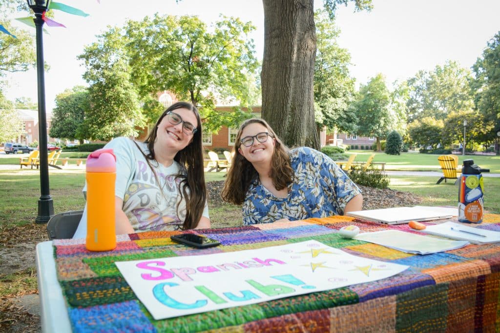 Two people sit at a table outdoors for the lively Spanish Club, with colorful signs and water bottles, surrounded by trees and green grass.