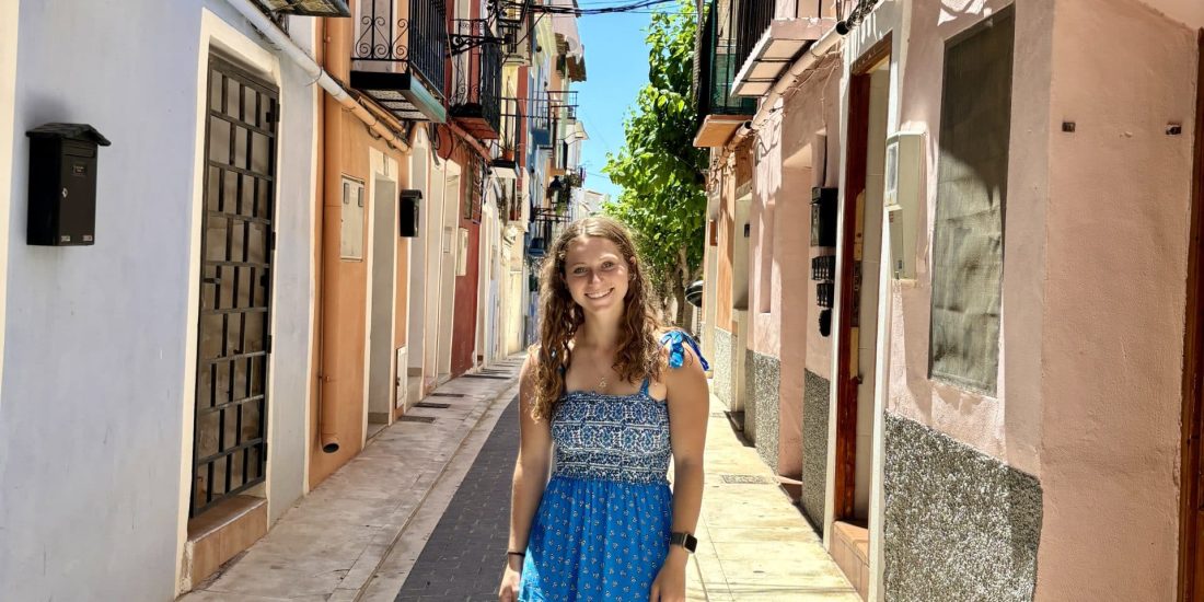 A young woman in a blue dress stands in the middle of a narrow street with colorful buildings and balconies on each side. The sky is clear and sunny.