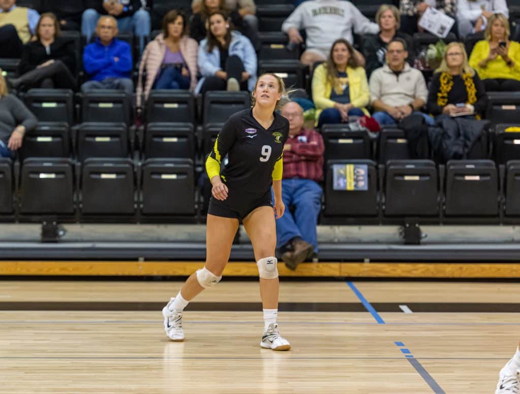 A volleyball player wearing a black jersey with the number 9 prepares on the court with spectators watching in the background.