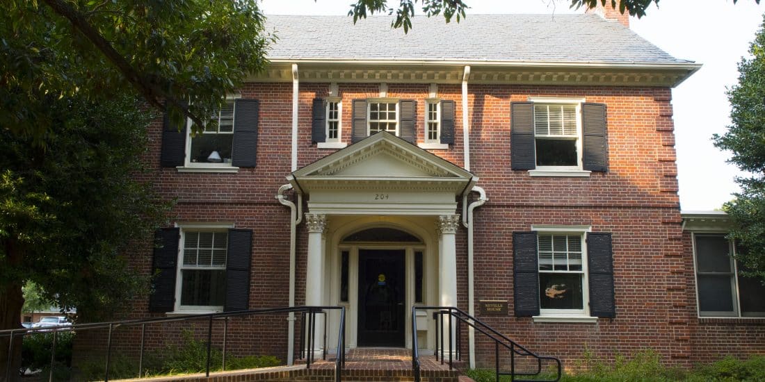 A two-story red brick building with black shutters, a small front porch, and a railed wheelchair ramp leading to the entrance. Trees and bushes surround the building.
