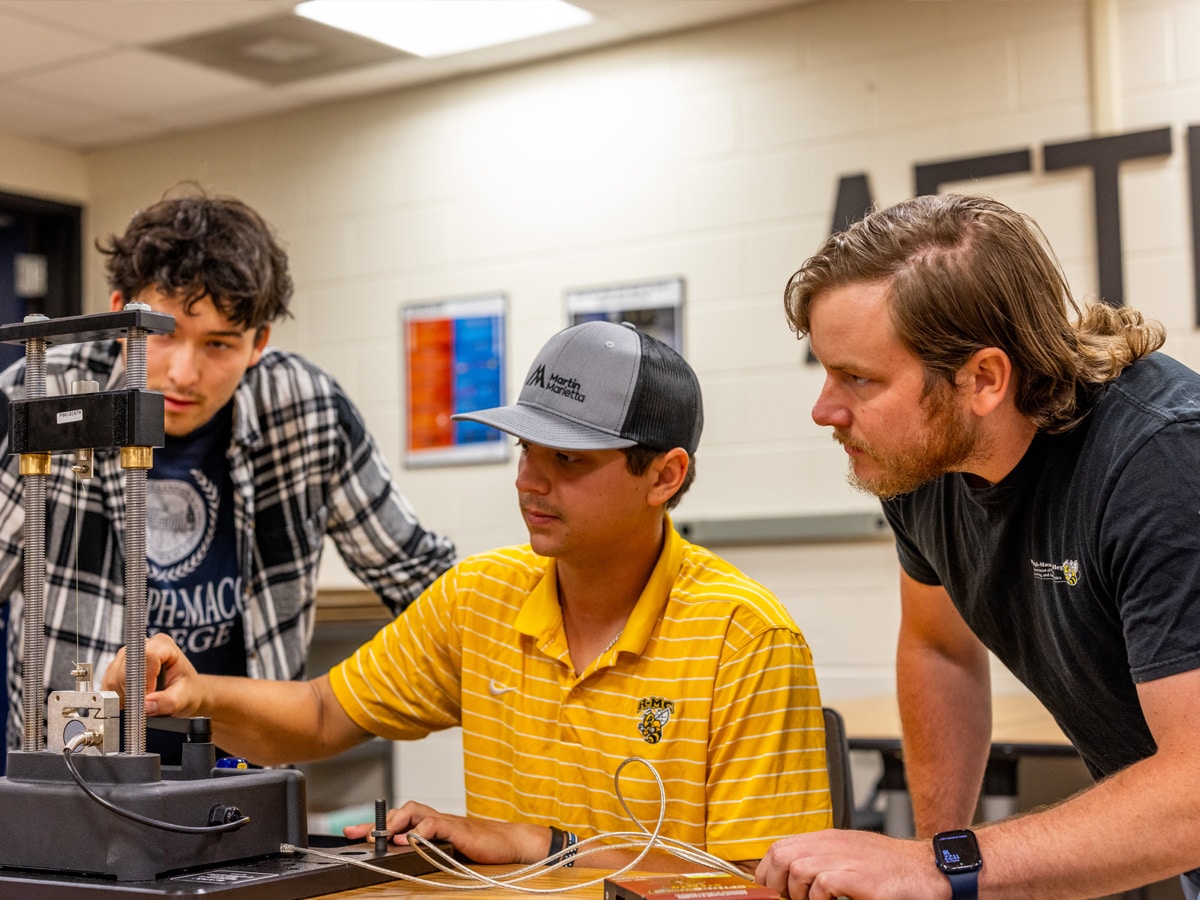 Three RMC students work together on an engineering project in a college classroom. One man adjusts the technical equipment, while the others look on attentively, making the space feel like a collaborative workshop at home.