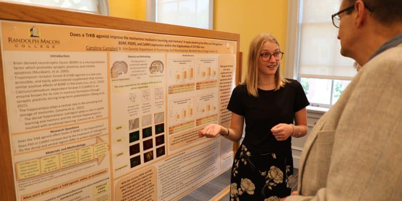 An RMC student stands in front of a research poster, explaining its contents to a man in a gray jacket. The poster displays graphs, text, and images under the title about a study on the role of TRKB signaling in memory.