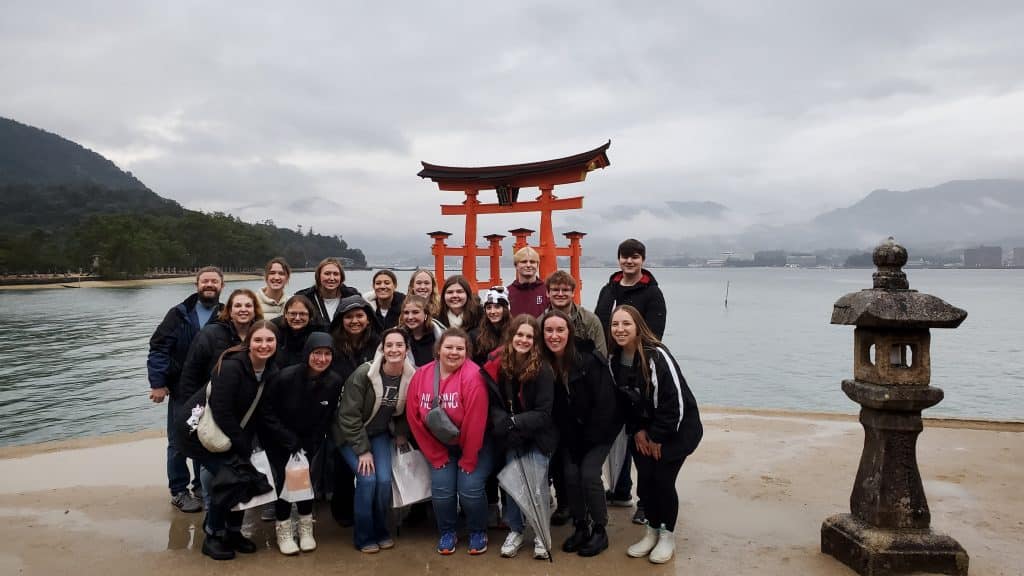 A group of people, including nursing students and professors, stand in front of the red torii gate at Itsukushima Shrine, Japan, on a cloudy day with a mountainous background.