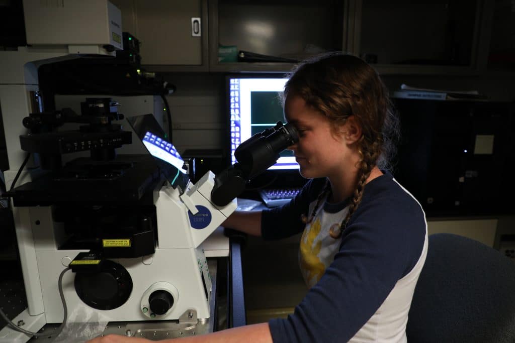 Person looking through a microscope in a laboratory setting, with a computer screen in the background displaying data.