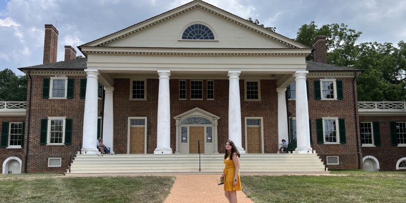 A person in a yellow dress stands on a pathway in front of a large two-story building with columns and a triangular pediment.
