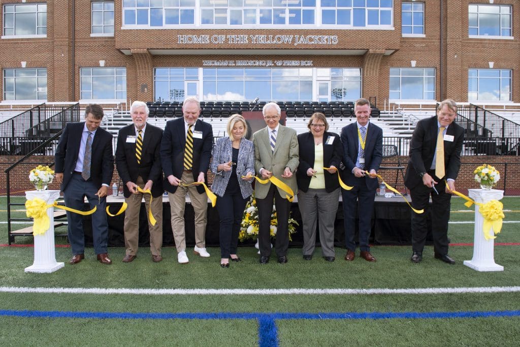 Eight college leaders and trustees stand in a row cutting a yellow ribbon during a ceremony in front of Duke Hall, a building labeled Home of the Yellow Jackets, commemorating the new facilities.