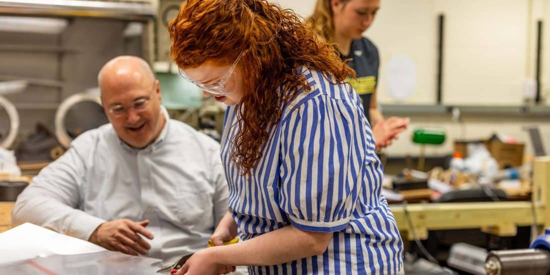 Three people collaborate around a workbench in a workshop. One person is seated and two are standing, all engaged in a project involving tools and materials.