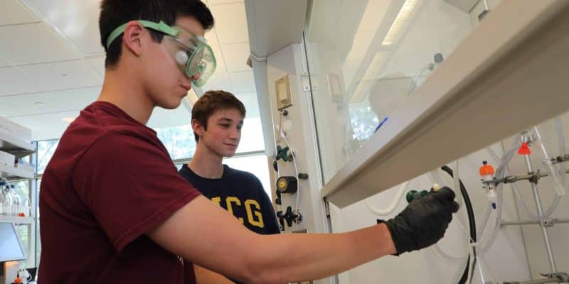 Two individuals wearing gloves work on an experiment in a laboratory. One operates equipment inside a fume hood, while the other observes. They wear safety glasses and casual attire. Various lab equipment is visible.