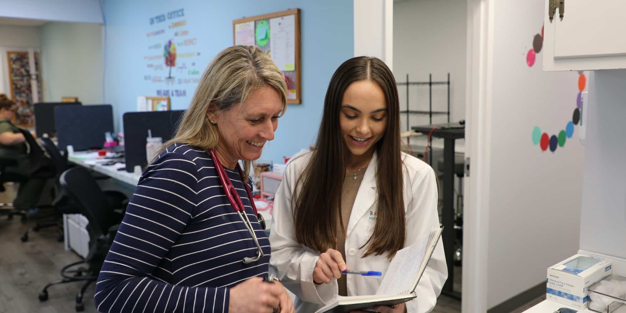 Two women, one in a striped top with a stethoscope and the other in a white lab coat, look at a notepad and smile in an office setting. The assistant appears to be helping the physician with some studies.
