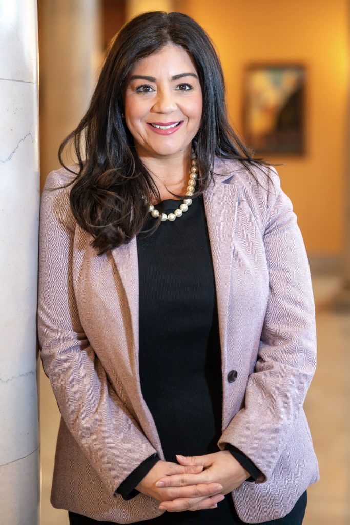 A person with long dark hair wearing a pink blazer over a black top stands indoors, smiling and posing with clasped hands in front of a column.