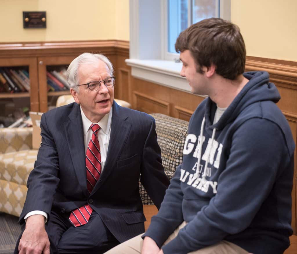 President Lindgren in a suit converses with a male student wearing a hoodie, seated