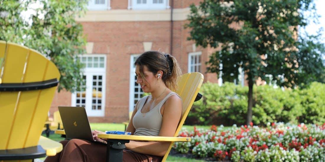 A person sits on a yellow chair outdoors, working on a laptop, with a brick building and a garden in the background, possibly searching for study tips or strategies.