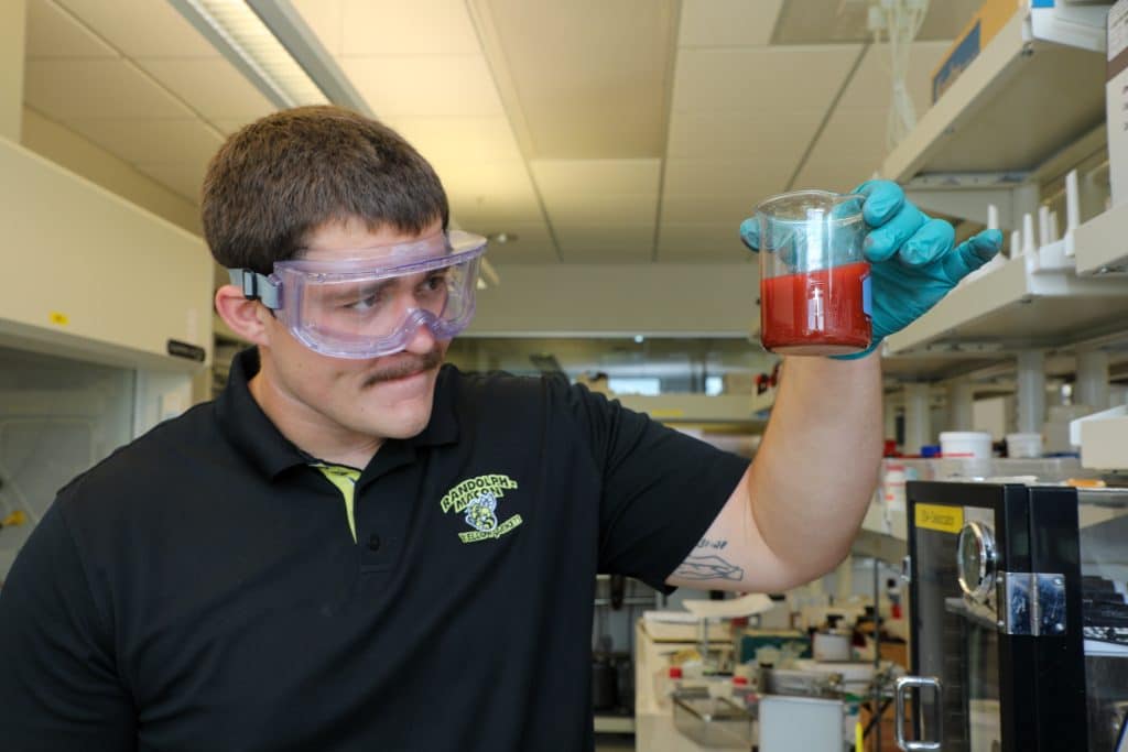 A person wearing safety goggles and gloves holds a beaker with a red liquid while standing in an RMC laboratory.