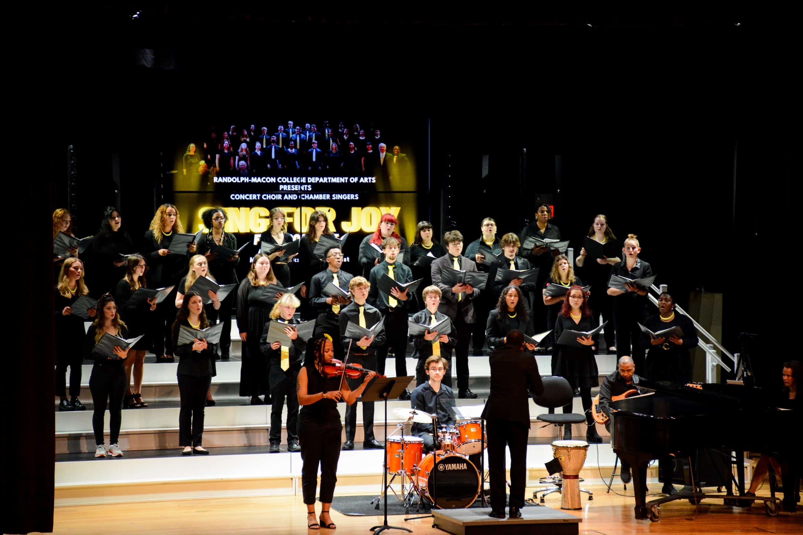 A choir performs on a staged platform with a conductor, musicians, and a sign reading "Randolph-Macon College Department of Arts presents Singing For Joy Fall 2022 Concert for R-MC Chamber Singers.