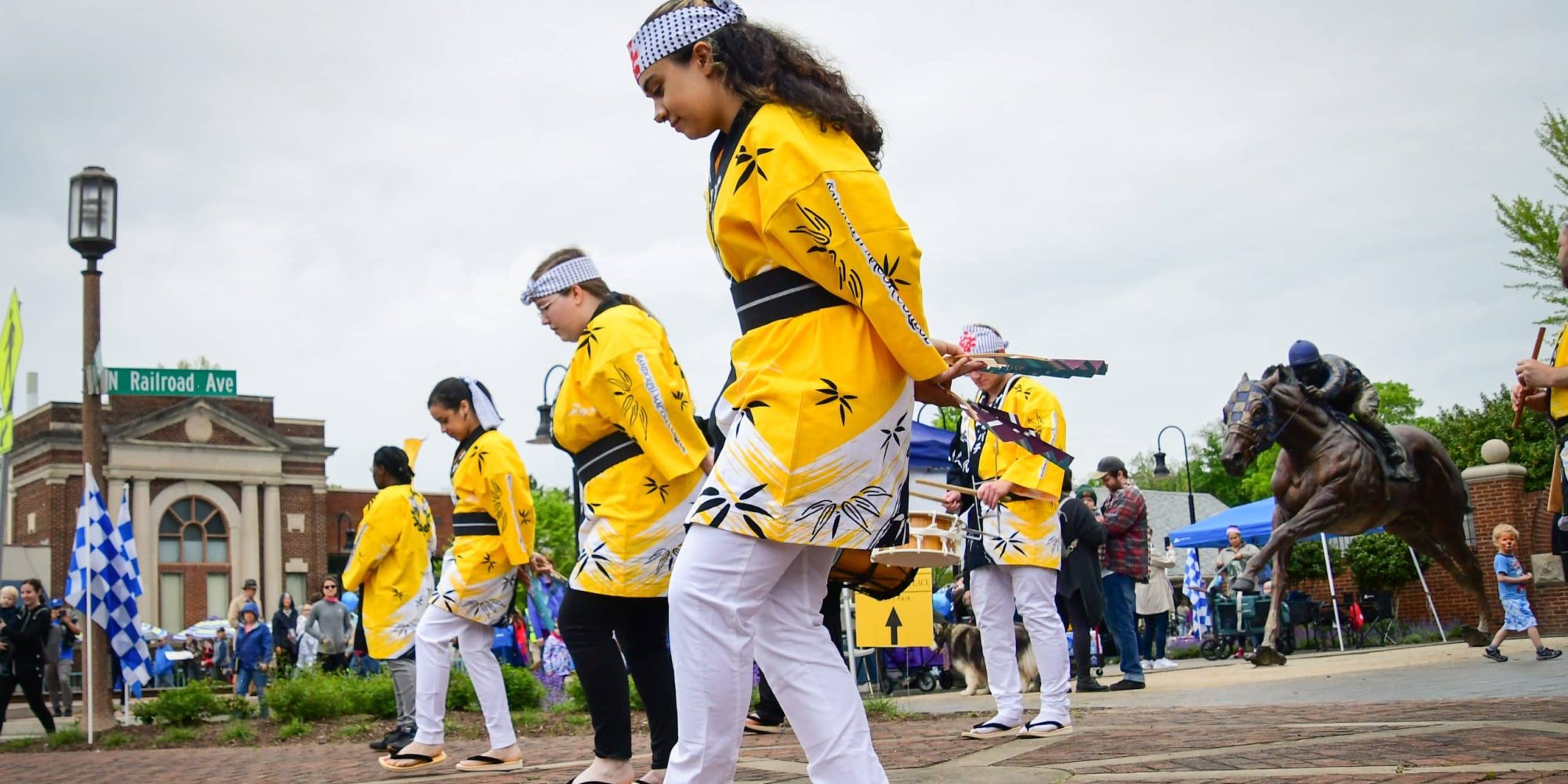 A group of people in yellow traditional outfits and tabi socks perform a dance on a brick-paved area, with spectators and various structures in the background.