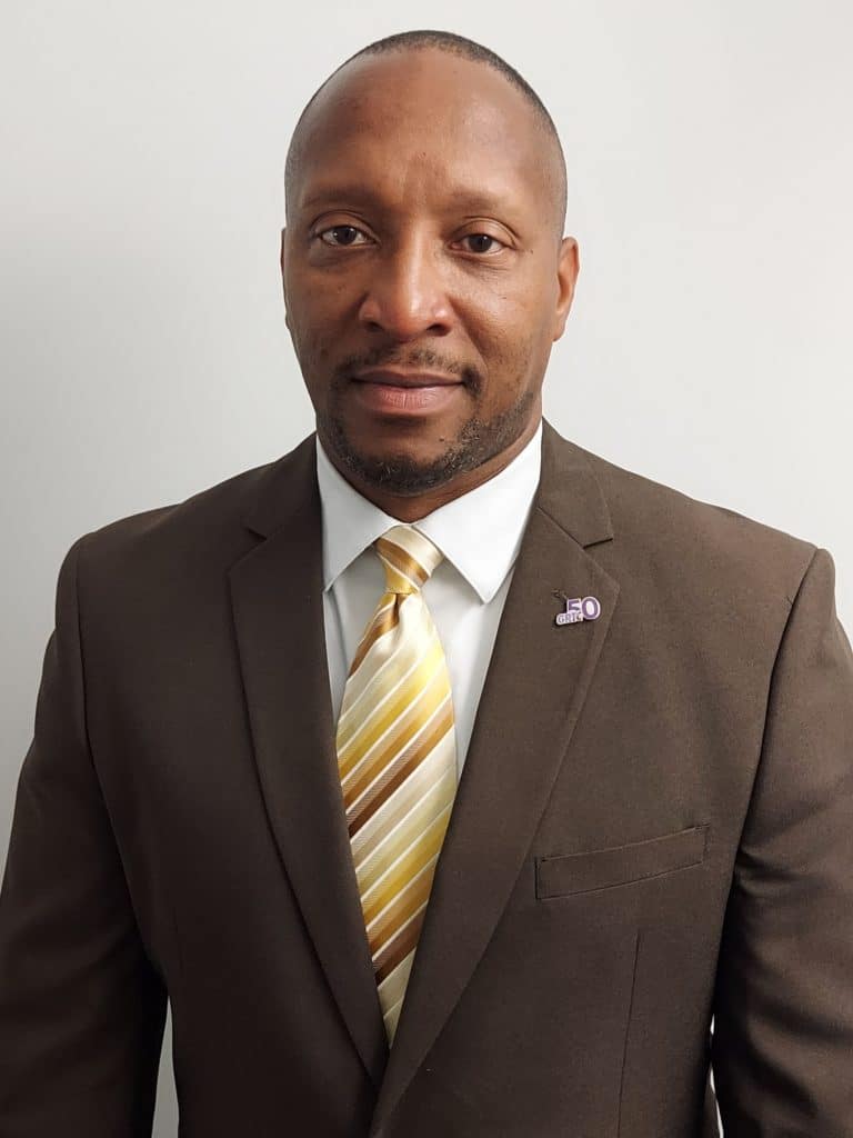 A man stands against a plain background wearing a brown suit, striped tie, and a lapel pin.