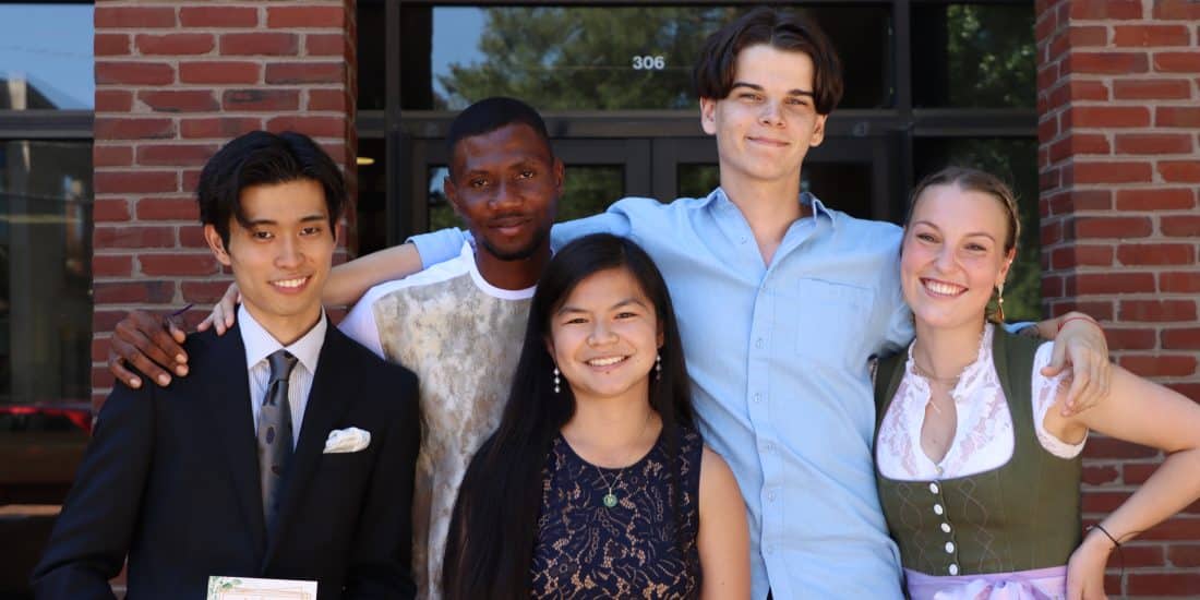 A group of five people stand close together in front of a brick building, smiling at the camera. The group includes two men in suits and three people in casual or semi-formal attire, perhaps gathering after a 日本語学習 session.