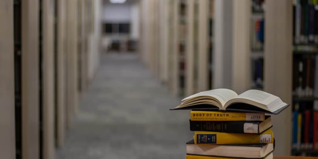 An open book sits atop a stack of four closed books on a table in the foreground, offering study tips, with a long, lit hallway flanked by bookshelves stretching into the background.