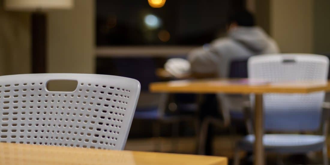 A white perforated chair is in the foreground of a dimly lit room with empty tables. In the background, a person in a grey hoodie sits at another table, perhaps contemplating study strategies.