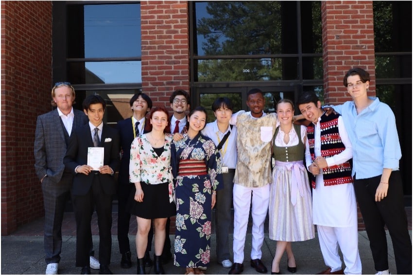 A group of 12 people stands together in front of a brick building. Dressed in a mix of formal and cultural attire, they smile for the photo. One person holds a sign with 日本語 written on it, adding an extra touch of cultural significance to the moment.