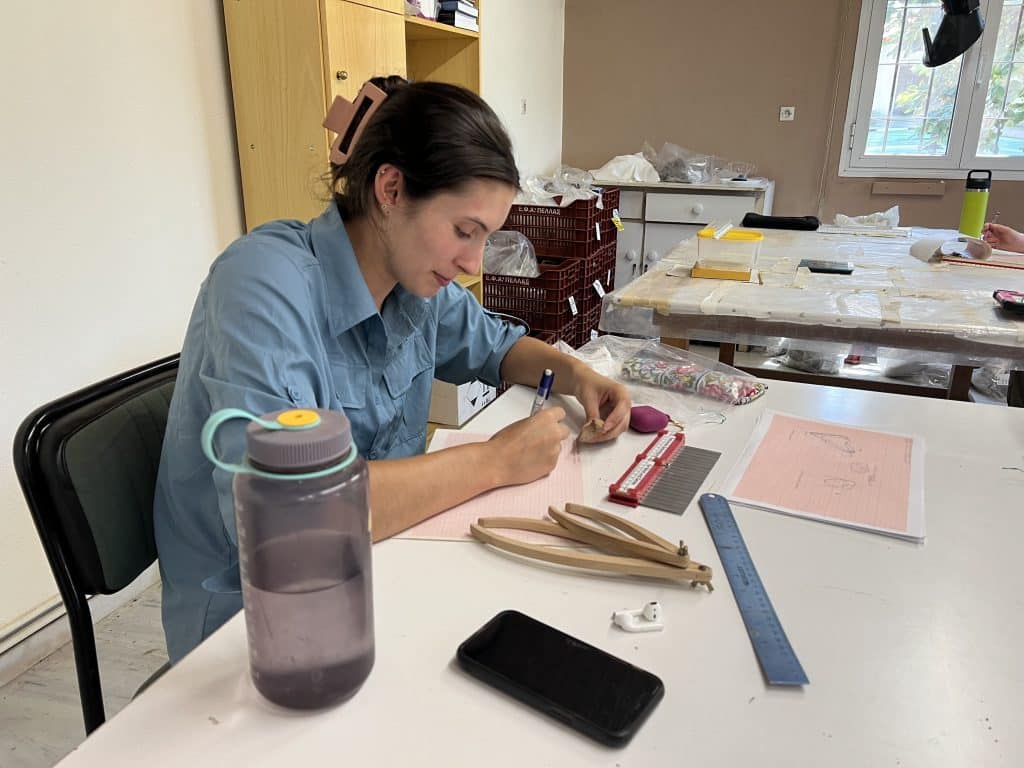 A person sits at a table writing on grid paper with a pen while holding an artifact. Nearby are a water bottle, smartphone, and various other items.