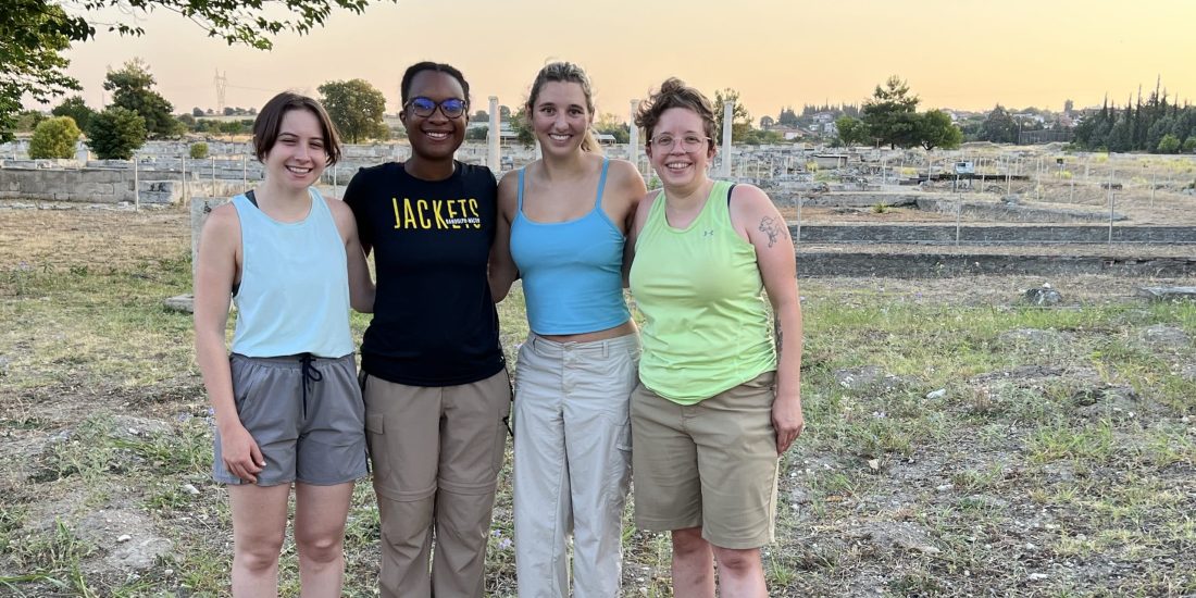 Four people stand together outdoors in a grassy area with a dig site in the background.