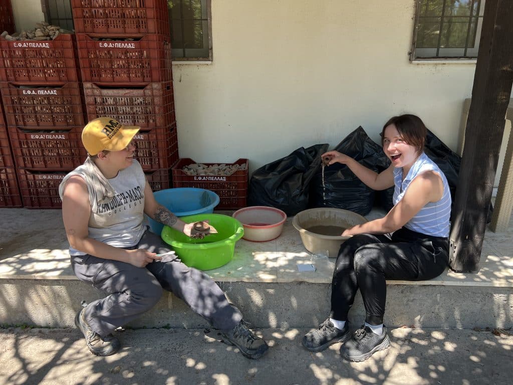 Two individuals sit on a step, sifting through artifacts in bowls. Behind them are stacked crates. One person holds an artifact in a green bowl, while the other reacts with surprise.