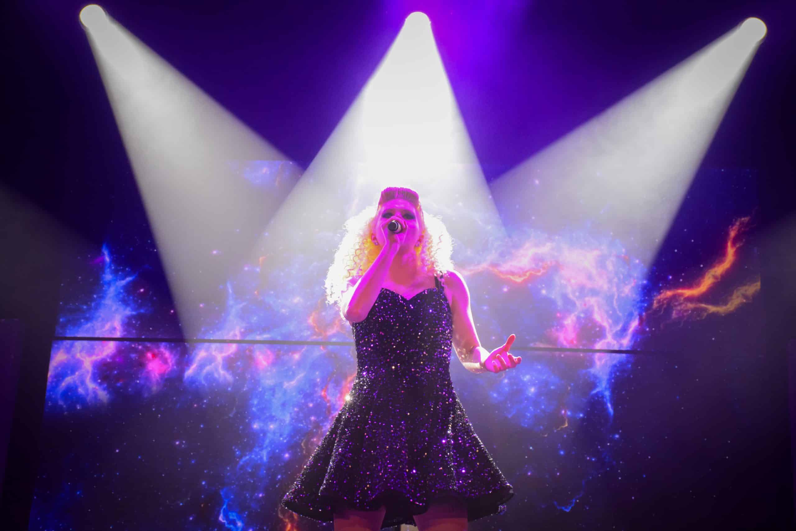 A singer performs on stage under bright spotlights, with a vibrant, colorful cosmic backdrop.