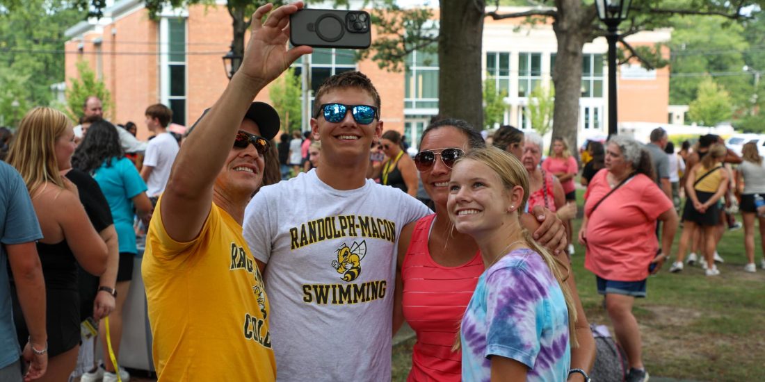 Four people stand closely together for a selfie, with one person holding a phone. They are outdoors with a crowd and buildings visible in the background.