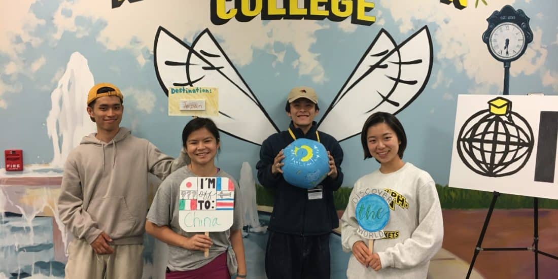 Four people stand in front of a Randolph-Macon College mural holding props related to travel, including a globe and a sign with the text "I'm going to China," with another sign displaying 日本語.