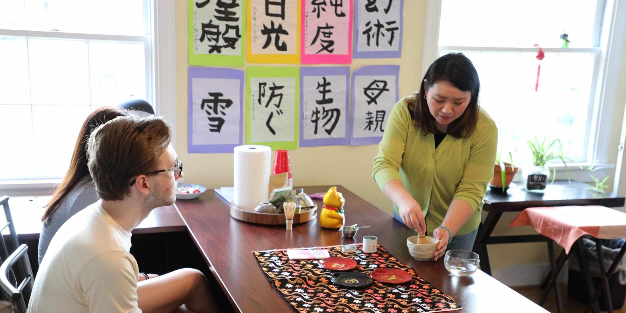 A woman, embodying the essence of Japanese Studies, demonstrates a traditional tea-making process to a seated group in a room adorned with colorful Japanese calligraphy on the walls.