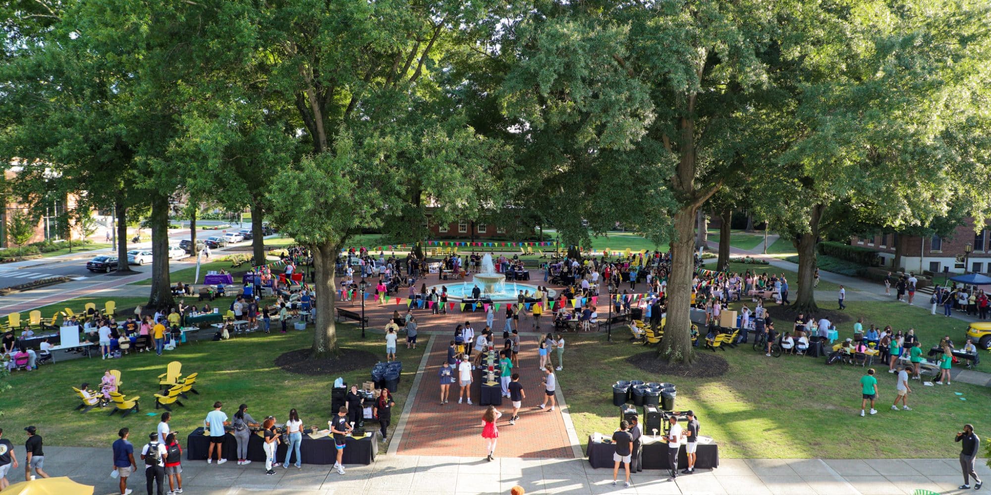 A large outdoor gathering with numerous people socializing, various activity booths, and food stations set up under tall trees, creating an atmosphere of student engagement as clubs promote their activities along the sidewalk dividing the area.