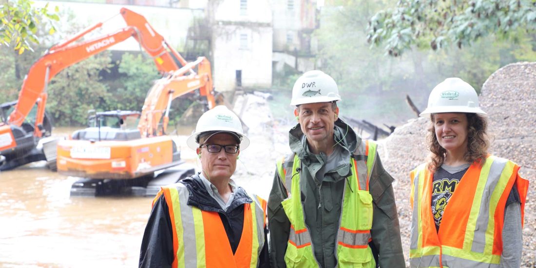Three people wearing safety gear stand by a construction site with two orange excavators near a river in the background.