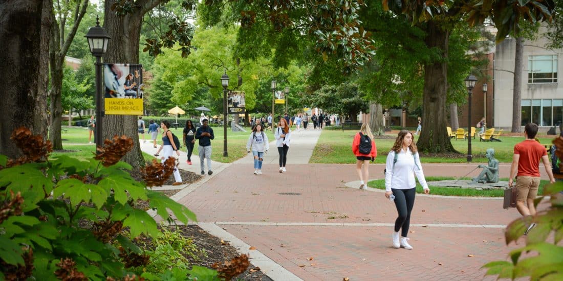 People walking along a tree-lined path on a college campus, with buildings and benches in the background.
