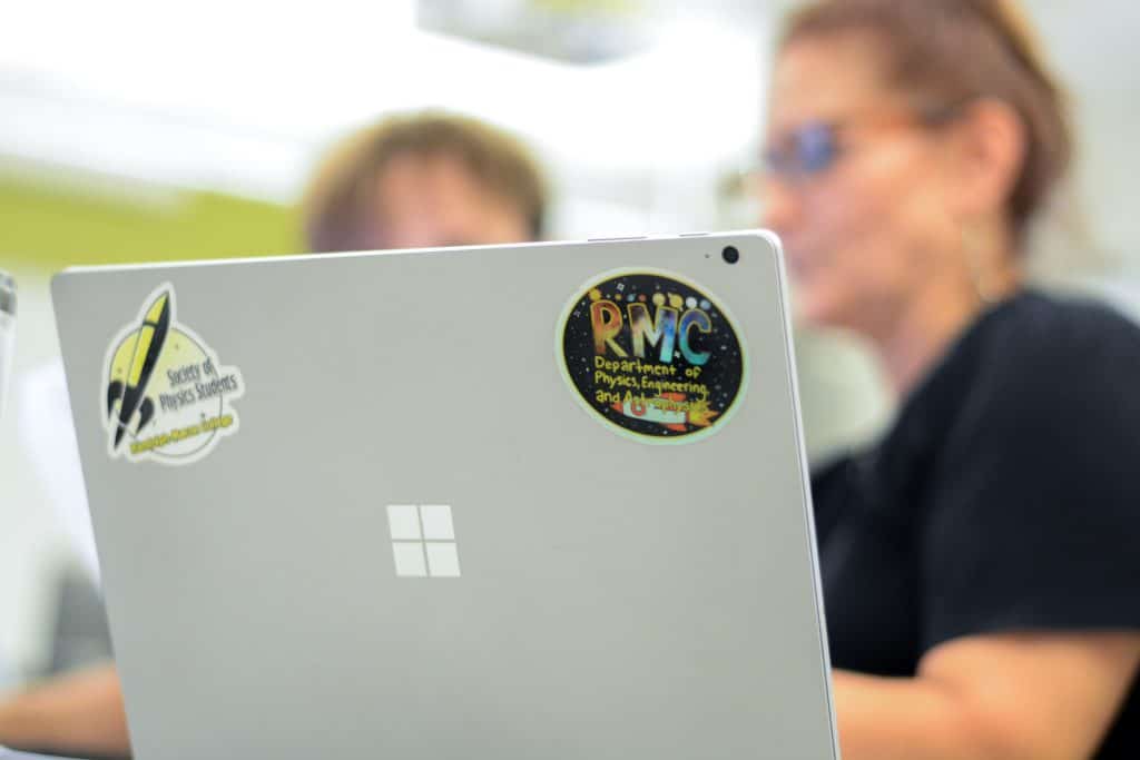 Two people collaborate on a Microsoft Surface laptop adorned with physics-themed stickers, their curiosity piqued by the secrets of the universe.