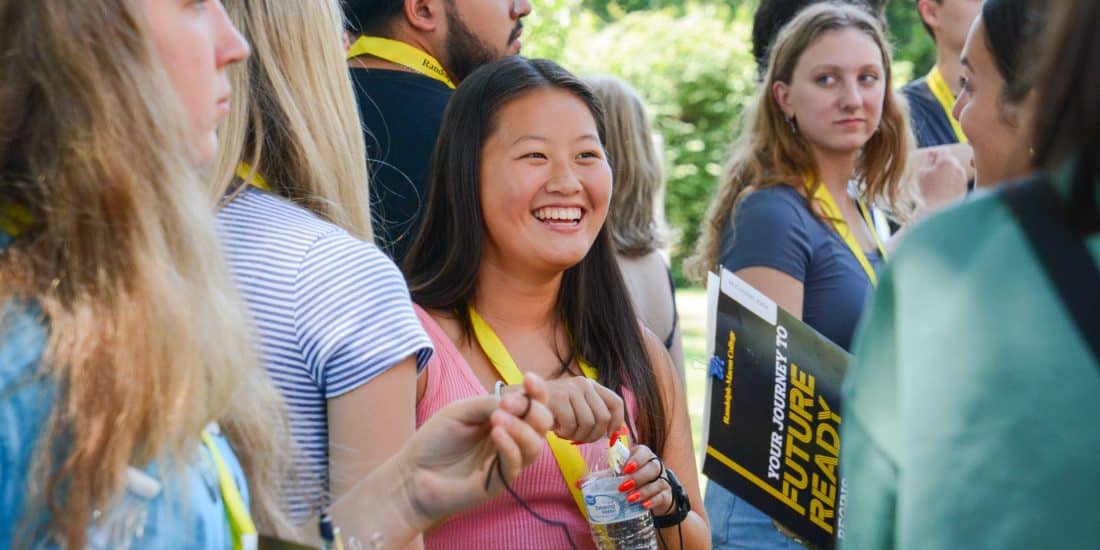 A group of people wearing yellow lanyards are gathered outdoors. A smiling woman in a pink top holds a water bottle and a booklet. Trees are visible in the background.