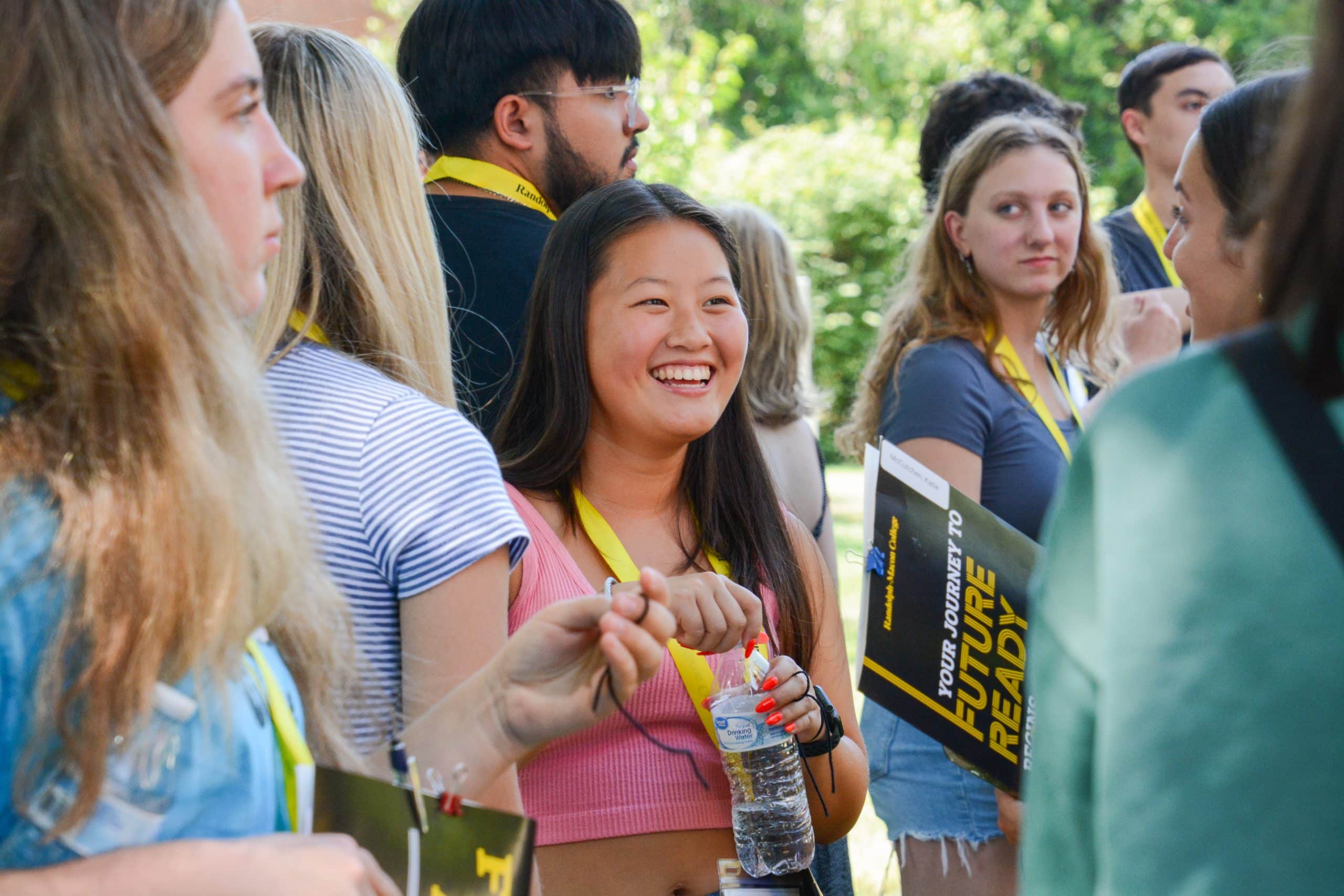 A group of people wearing yellow lanyards are gathered outdoors. A smiling woman in a pink top holds a water bottle and a booklet. Trees are visible in the background.